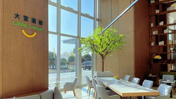 A modern lobby with a large glass window, a wooden wall with signage, a seating area with tables and chairs, and a bookshelf.