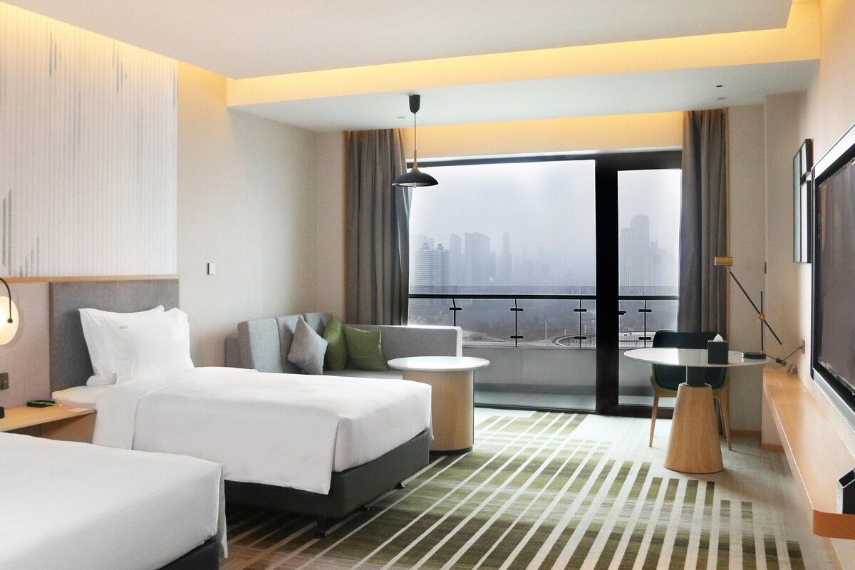 Ein Hotelzimmer mit zwei Betten, einem Sofa, einem kleinen Tisch und einem Balkon mit Blick auf die Stadt.