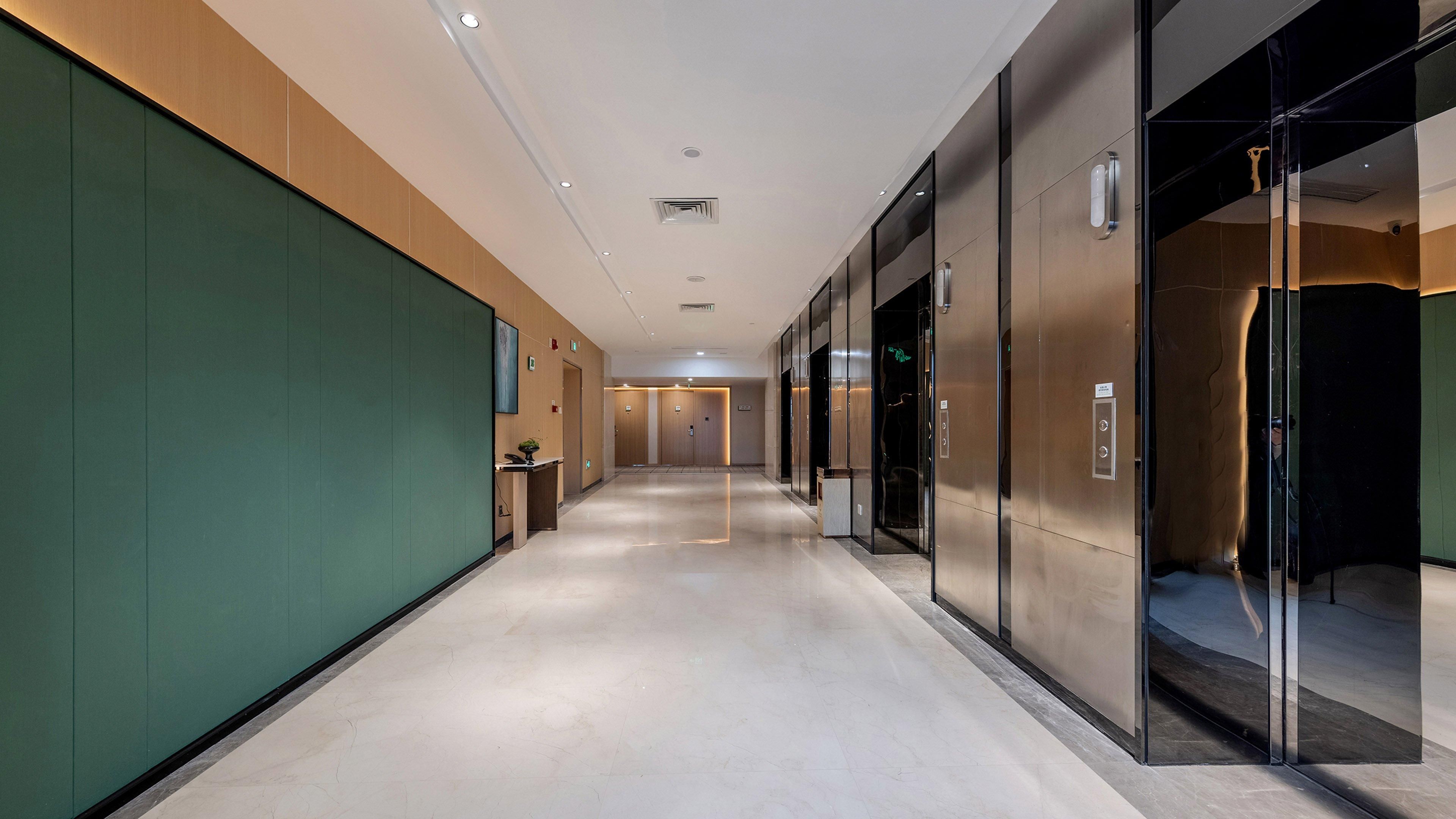 A modern hotel corridor with a long hallway, elevators, and a reception desk.