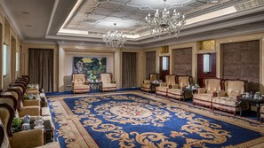 Uma sala de reuniões formal com um grande tapete azul, cadeiras ornamentadas, um candelabro e um quadro na parede.