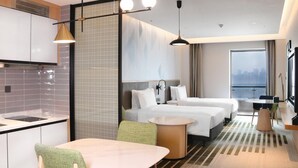 Una habitación de hotel moderna con zona de cocina, dos camas, una mesa de comedor y un amplio ventanal con vista a la ciudad.