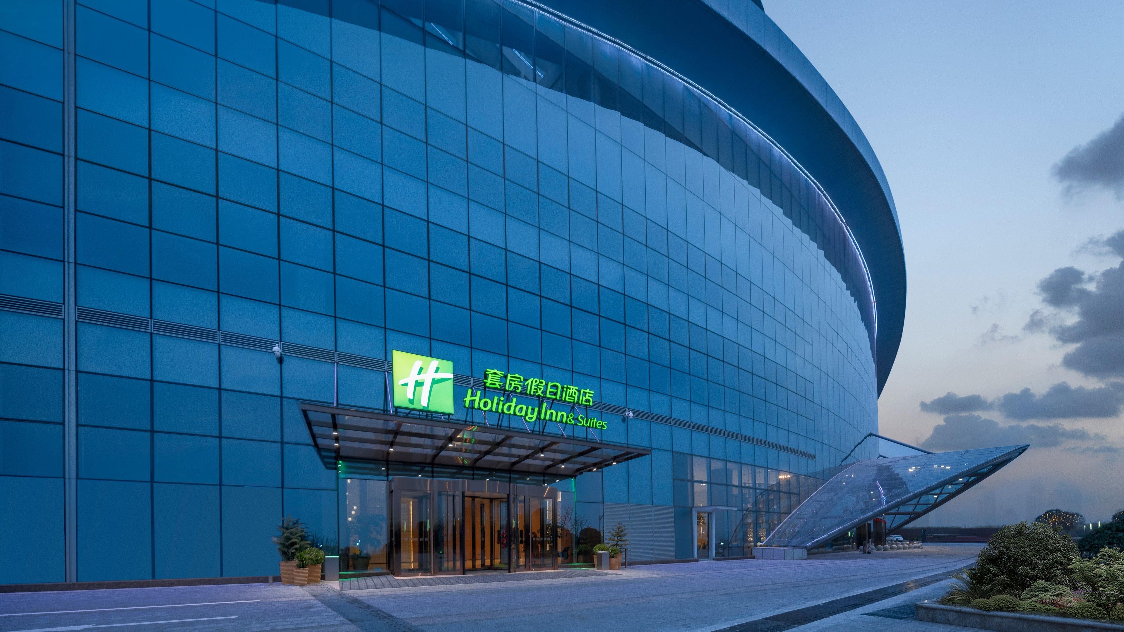 A modern building with a glass facade and a curved entrance. There is a sign for 'Holiday Inn & Suites' in English and another language.