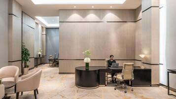 A modern reception area with a curved desk, a laptop, and a person seated. There are chairs, a potted plant, and a decorative vase.
