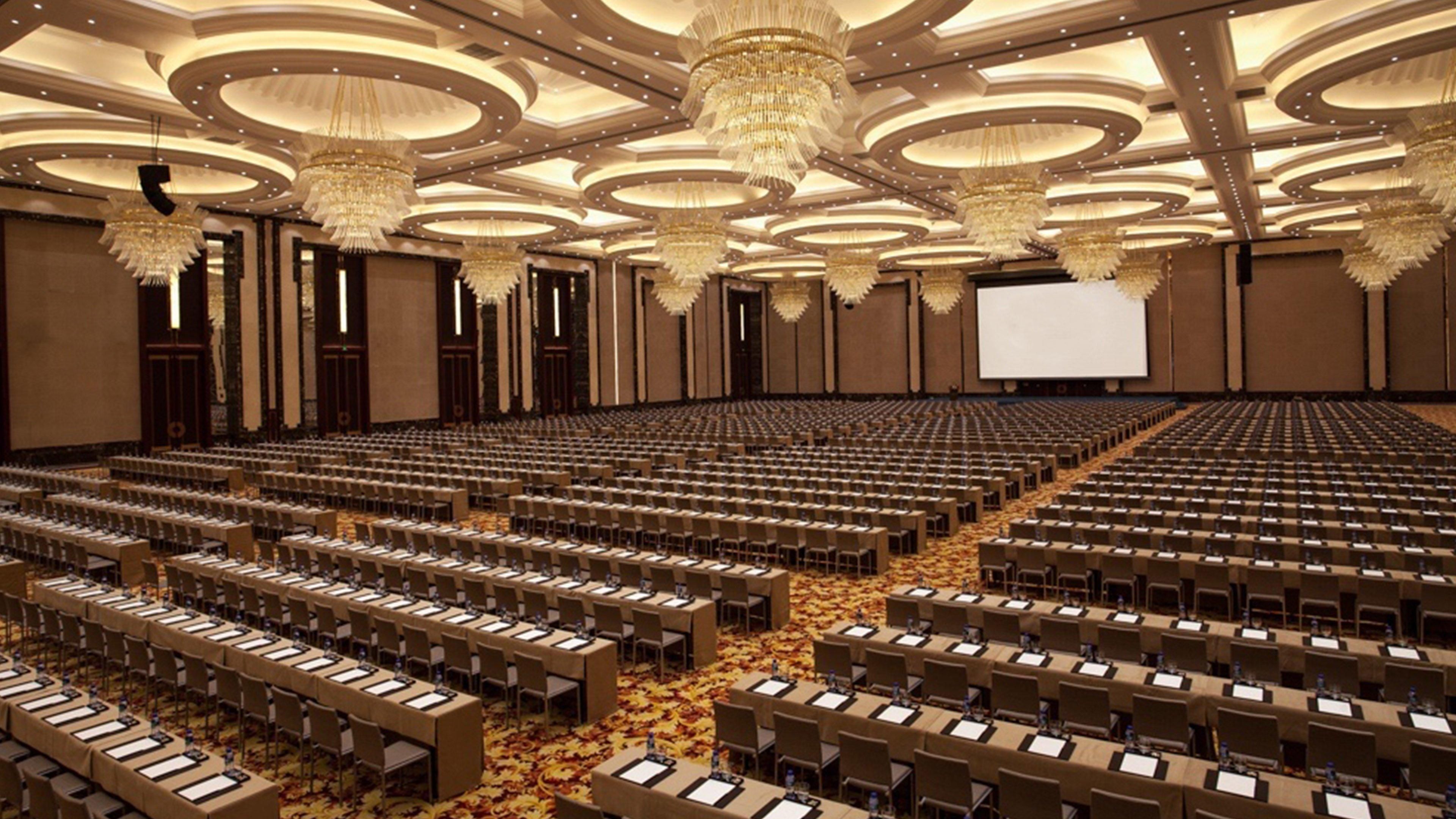 Un salón de conferencias amplio con filas de mesas y sillas, una pantalla grande al frente y candelabros decorativos.