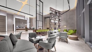 Eine moderne Lobby mit grauen Sofas, runden Tischen und grünen Stühlen. Eine Rezeption mit Computer und Pflanze ist vorhanden.