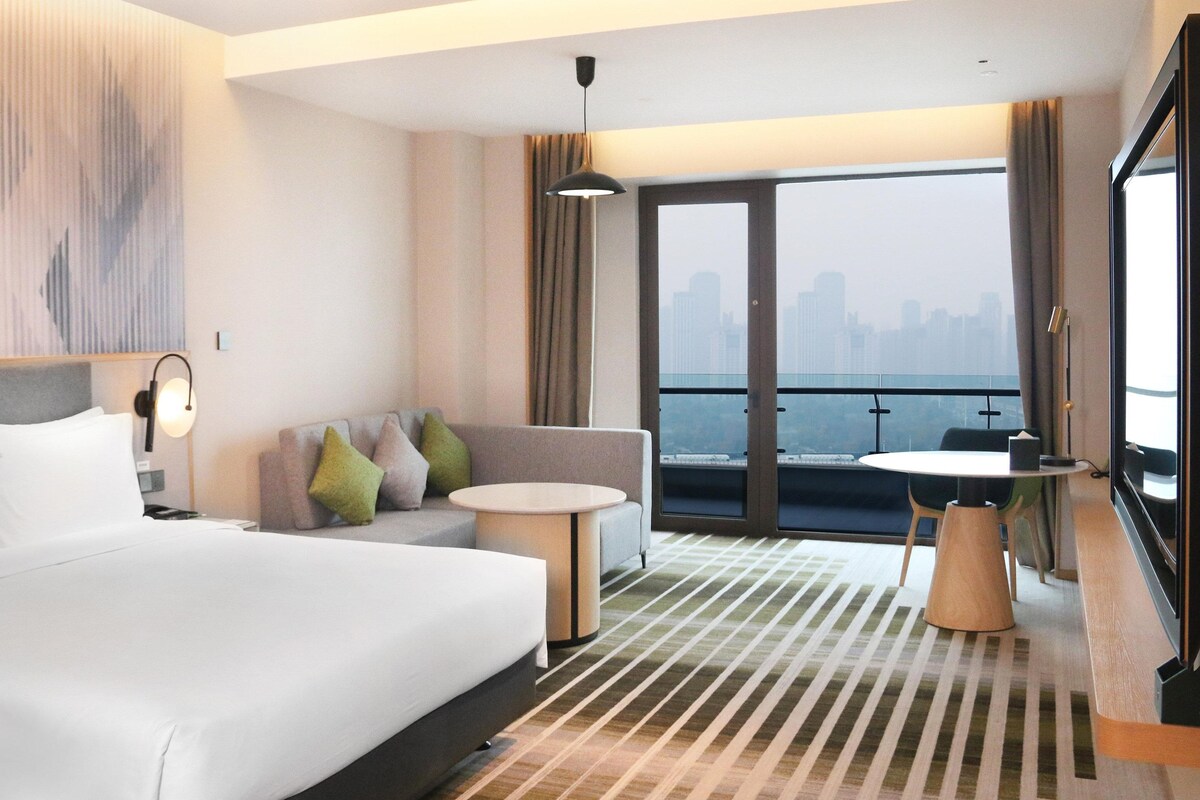 Ein Hotelzimmer mit einem großen Bett, einer Couch, einem kleinen Tisch und einem Balkon mit Blick auf die Stadt.