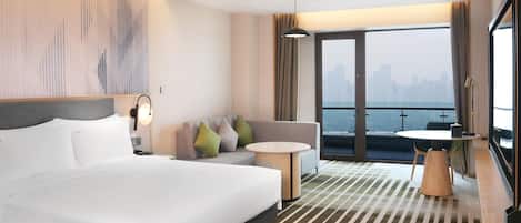 A hotel room with a large bed, a sofa, a small table, and a balcony with a view of the city.