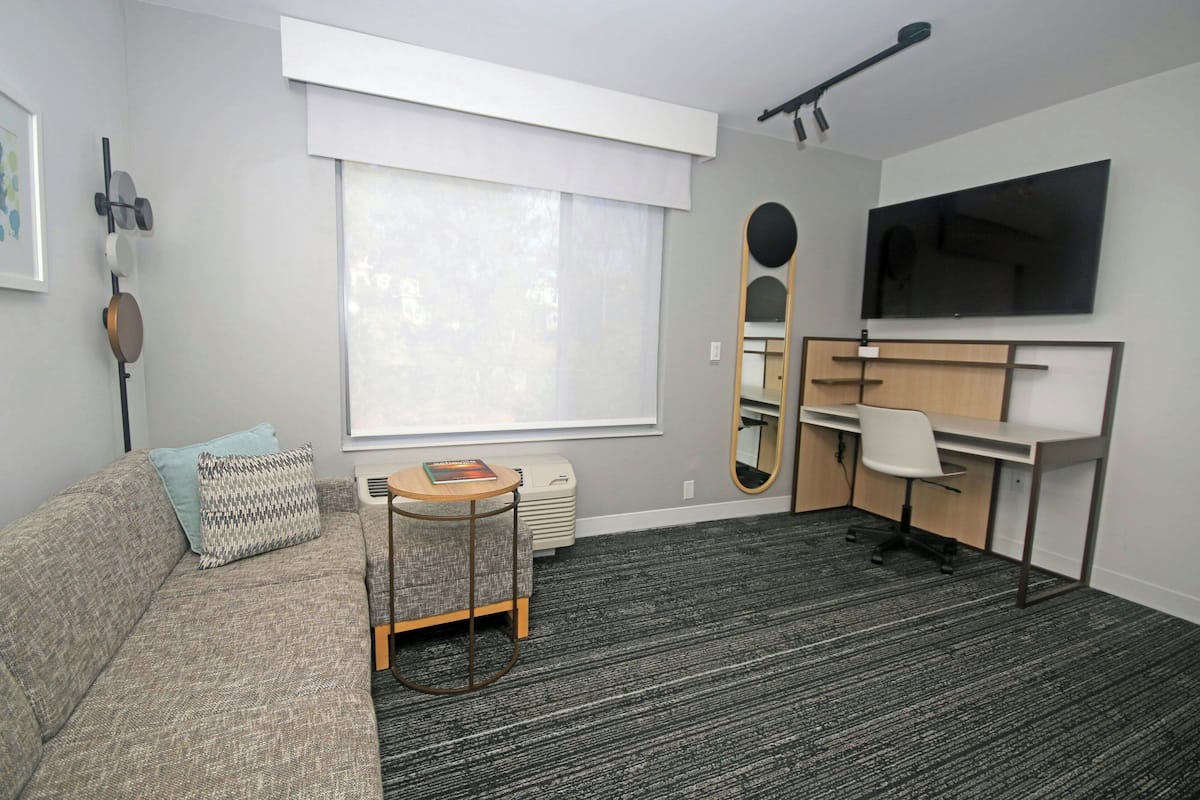A modern hotel room with a grey sofa, a small round table, a desk with a chair, a large mirror, and a flat-screen TV mounted on the wall.