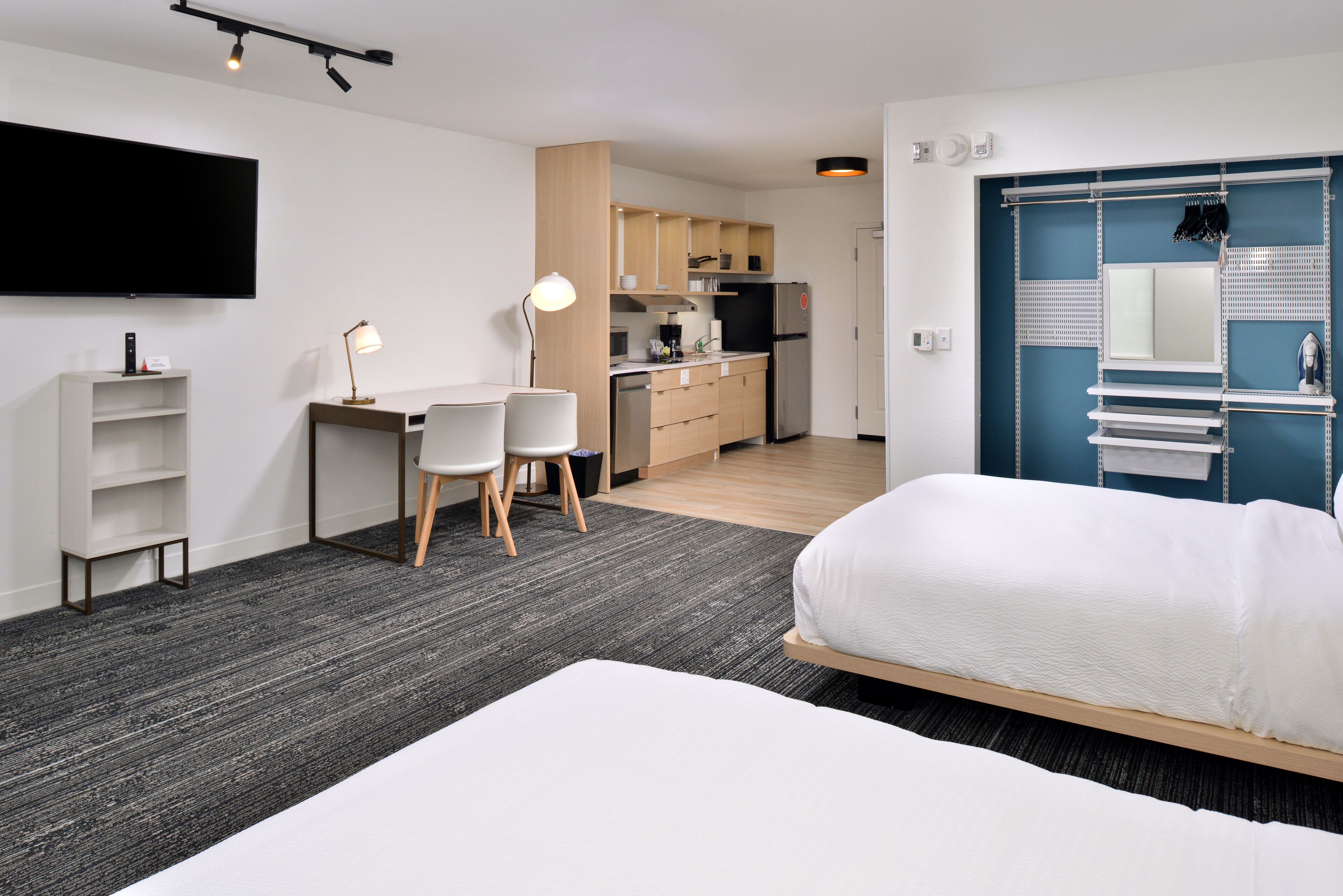 A modern hotel room with a large bed, a desk with two chairs, a kitchenette, and a TV mounted on the wall.
