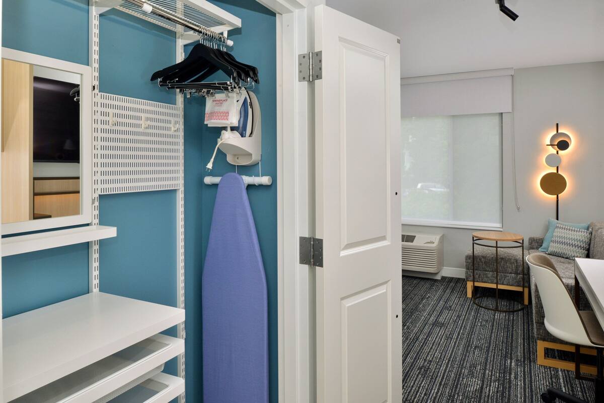 A hotel room with a blue wall, white door, and a small round table with a chair. There is a shelf with a blue towel and a hanging rack with clothes.