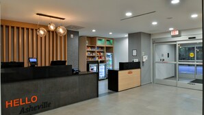 A reception area with a 'HELLO Asheville' sign, a reception desk, a vending machine, a refrigerator stocked with beverages, and an exit sign. - TownePlace Suites by Marriott Asheville Downtown (Asheville)