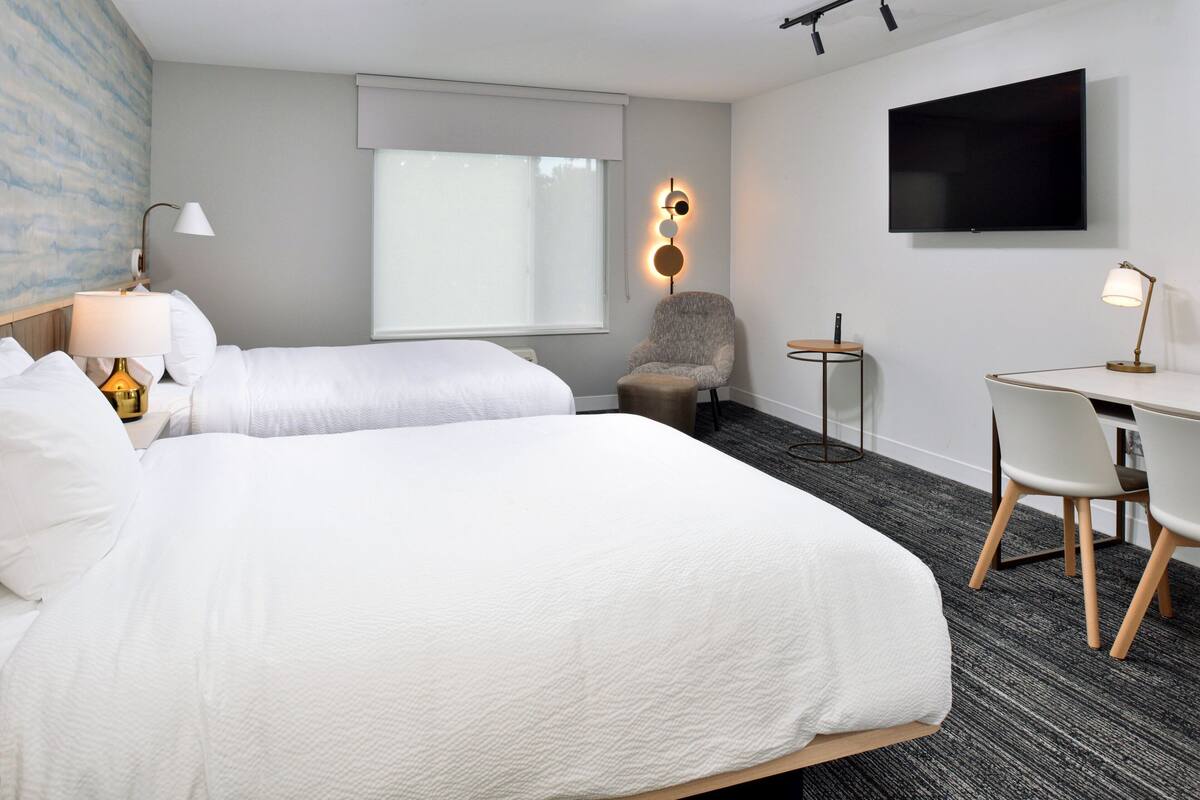 A hotel room with two beds, a desk, a chair, a TV, and a lamp.