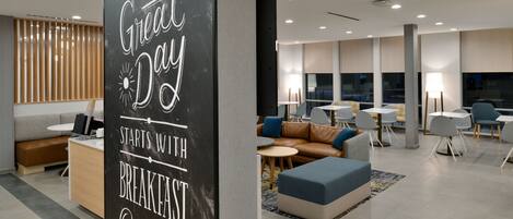 A modern hotel lobby with a black sign reading "A Great Day Starts with Breakfast." There are seating areas with sofas and tables, and a large window providing natural light.