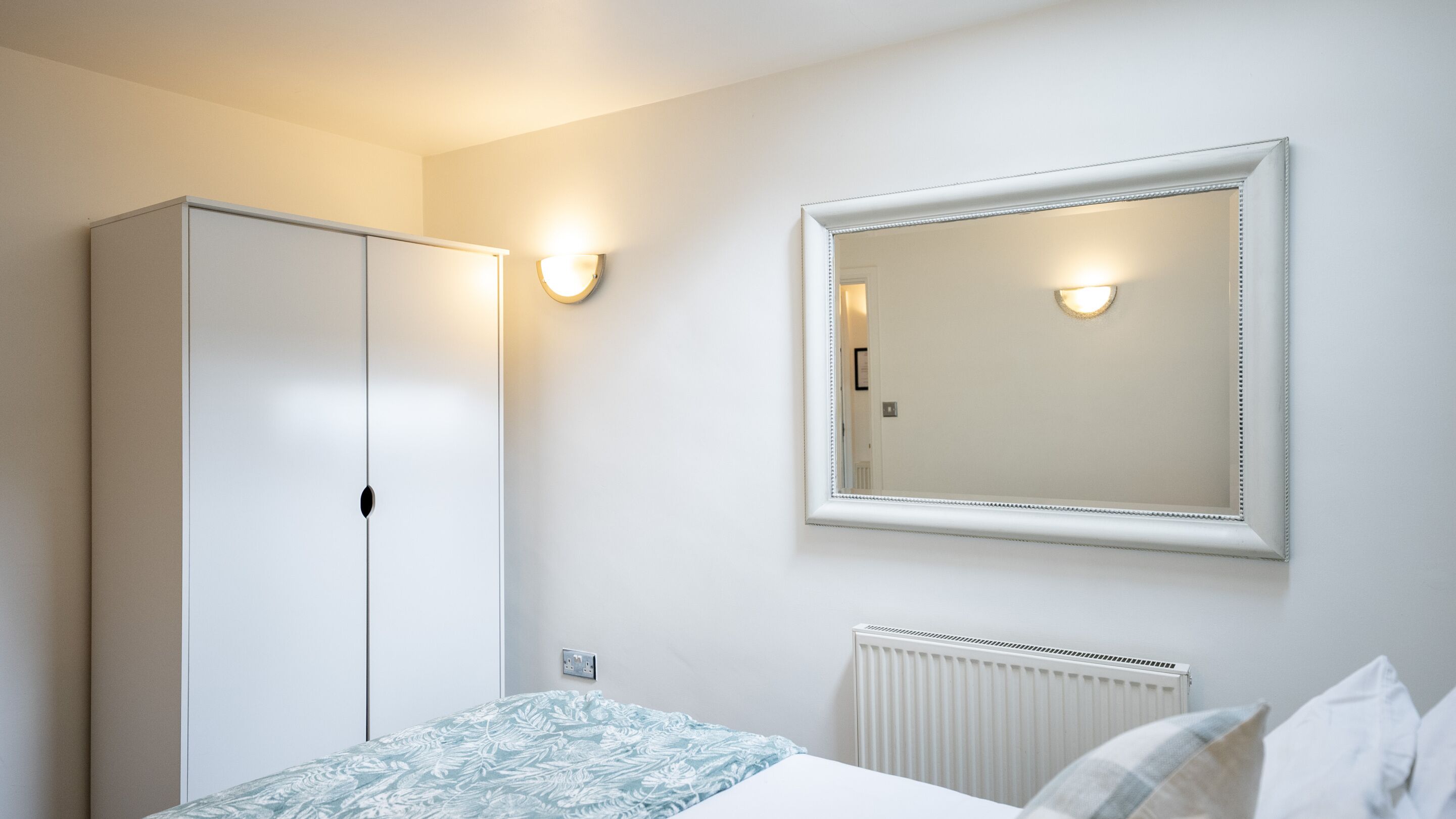 A bedroom with a wardrobe, a bed with a patterned bedspread, a wall-mounted mirror, and a radiator.