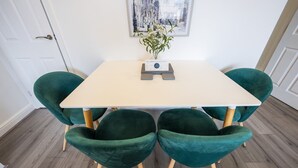 Un comedor con una mesa blanca, cuatro sillas tapizadas de verde y un florero con flores sobre la mesa.