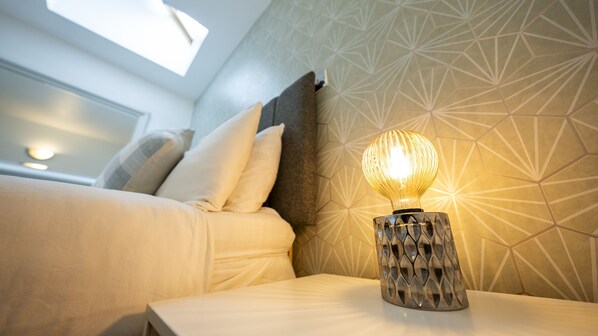 A neatly made bed with pillows, a bedside table with a decorative lamp, and a geometric patterned wall. - Modern Townhouse Apartment in Stratford Upon Avon With Wifi & Netflix (Stratford-upon-Avon)