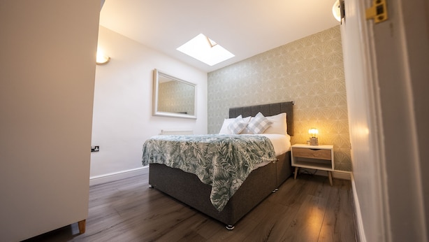 Un dormitorio con cama, mesita de noche, espejo en la pared y tragaluz.