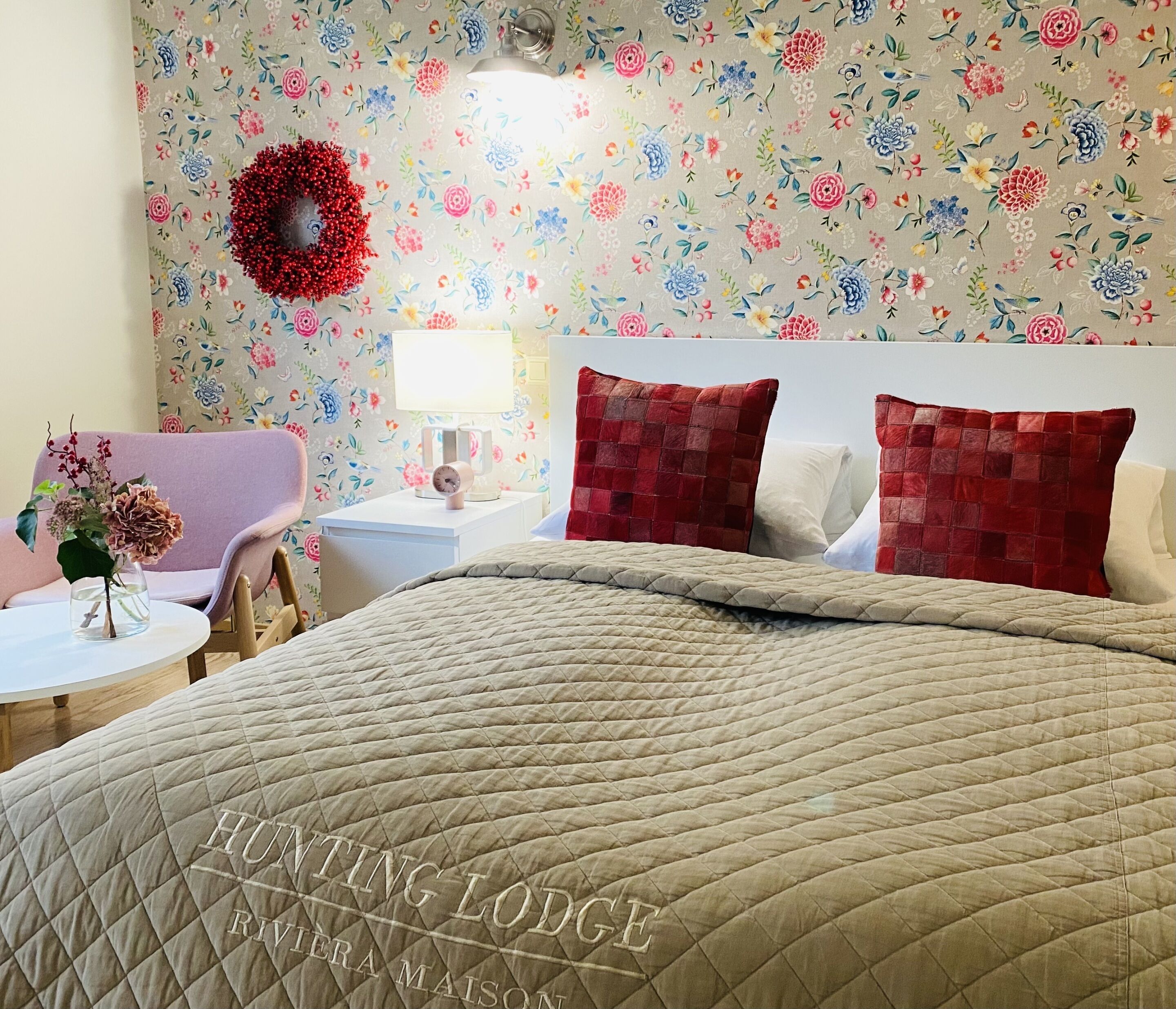 Une chambre d’hôtel avec un papier peint à motifs floraux, un lit doté d’une tête de lit matelassée, une table de chevet avec une lampe et une guirlande rouge accrochée au mur.