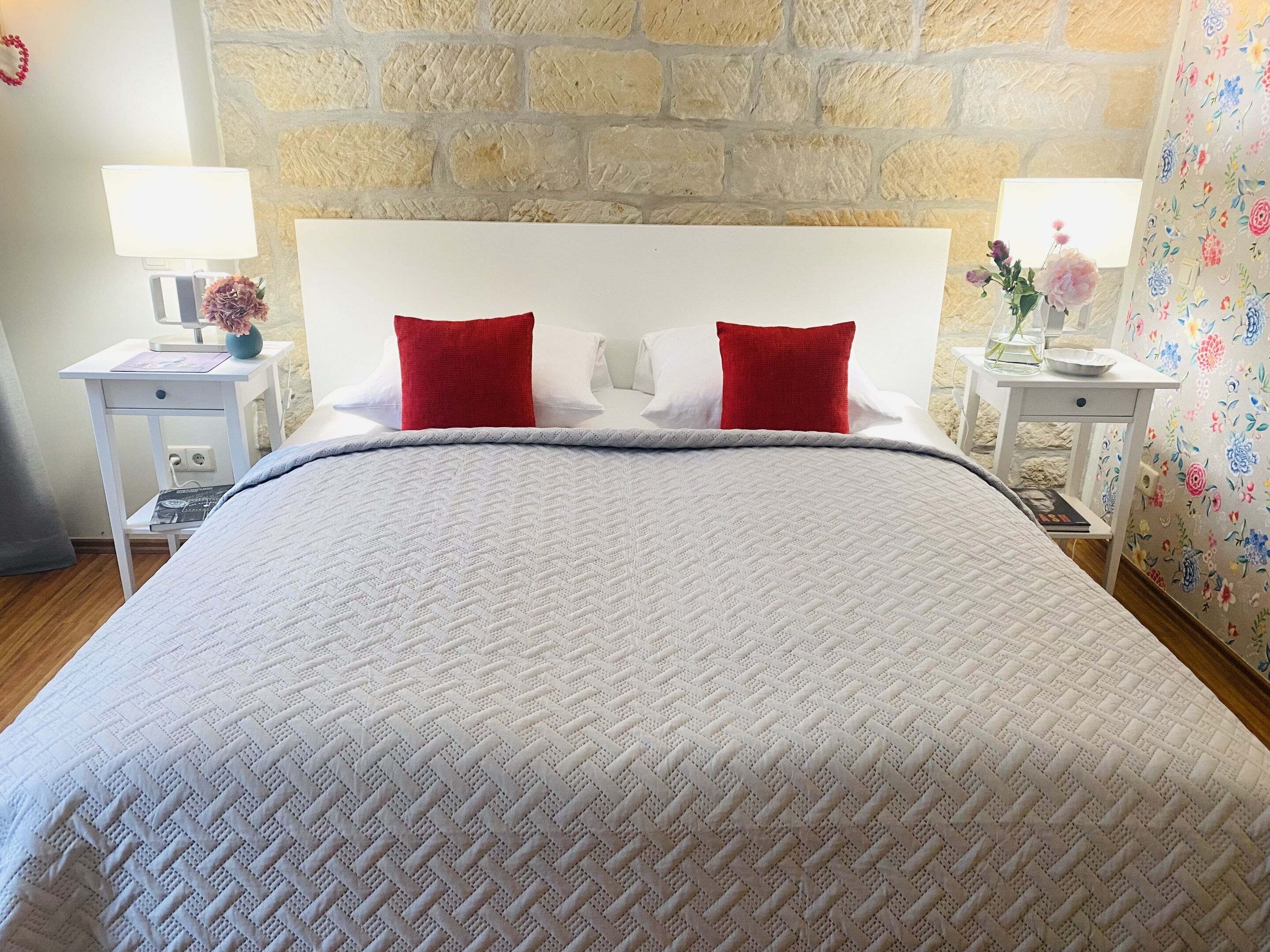 A neatly made bed with a white quilt and two red pillows, a white nightstand with a lamp and a vase of flowers, and a floral-patterned curtain.