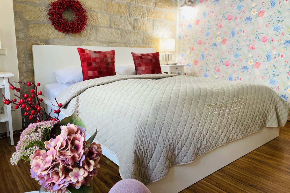 Un dormitorio con pared de piedra, papel tapiz floral, cama con colcha acolchada y una corona roja en la pared.
