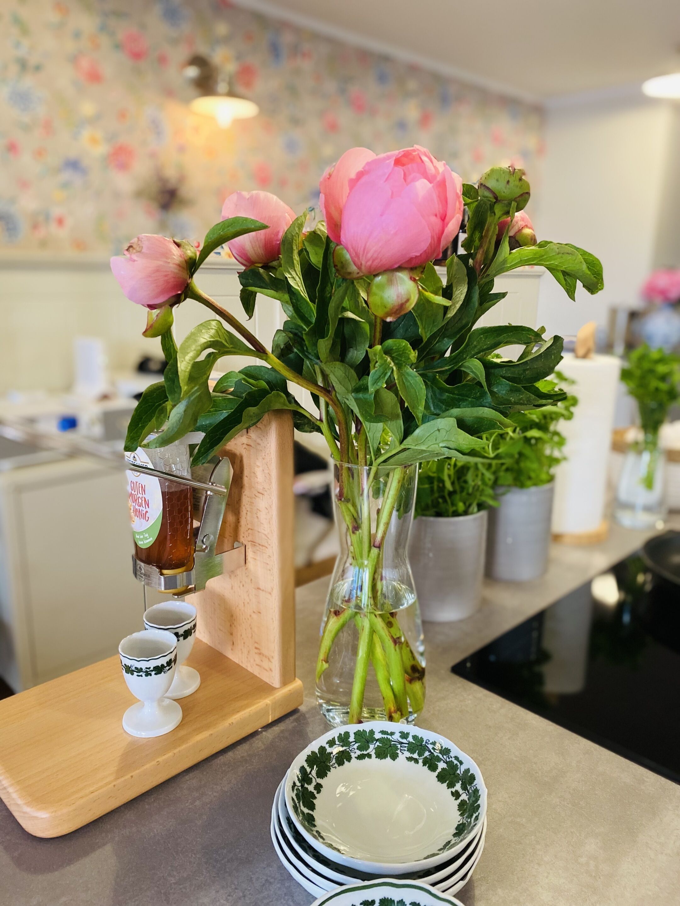 Eine Küchenarbeitsplatte mit einer Vase voller pinkfarbener Blumen, einer Kaffeemaschine und gestapelten weißen Tellern mit grünen Mustern.