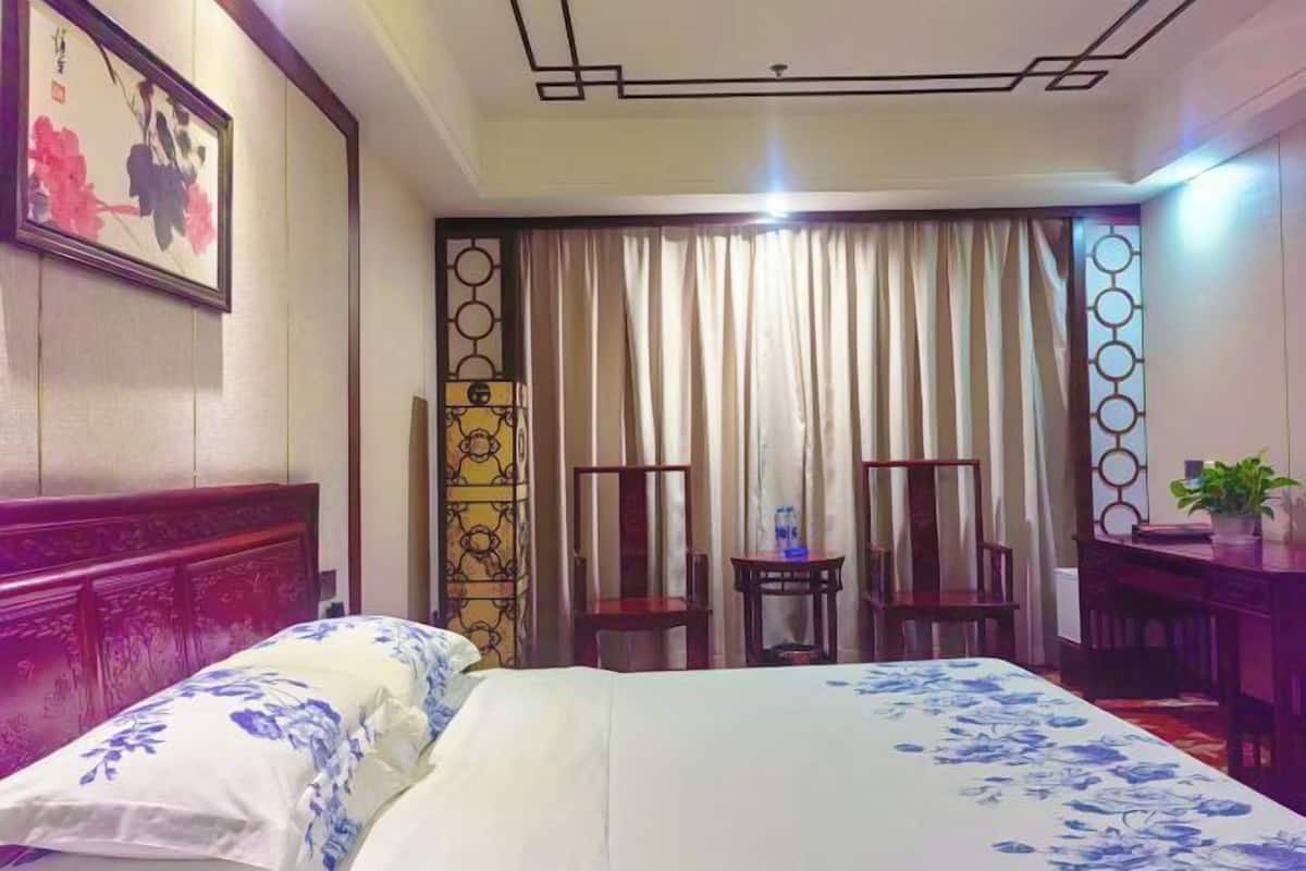 A hotel room with a bed, two chairs, a small table, a potted plant, and a decorative wall panel.