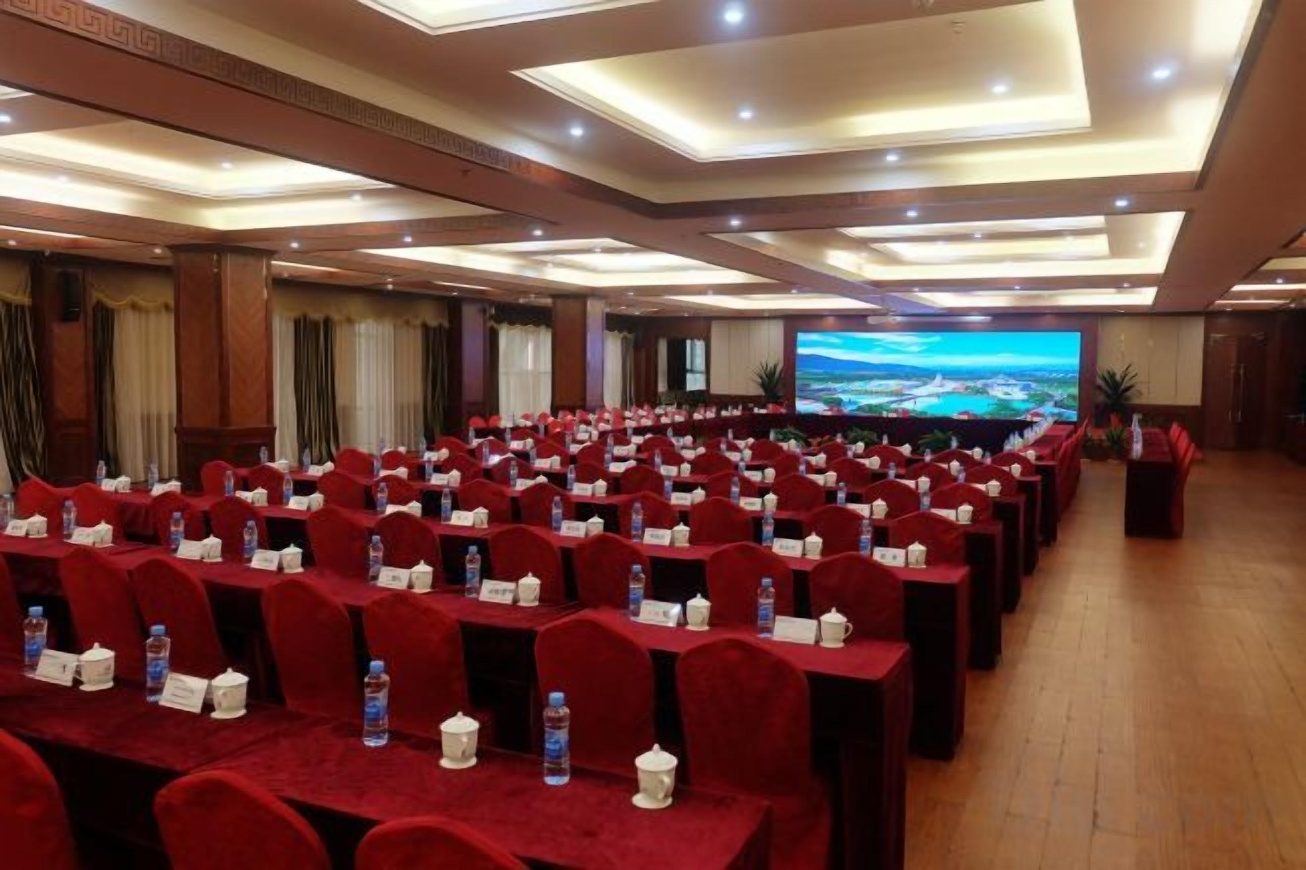 A conference room with rows of tables and chairs, a large screen at the front, and a view of a landscape through the windows.