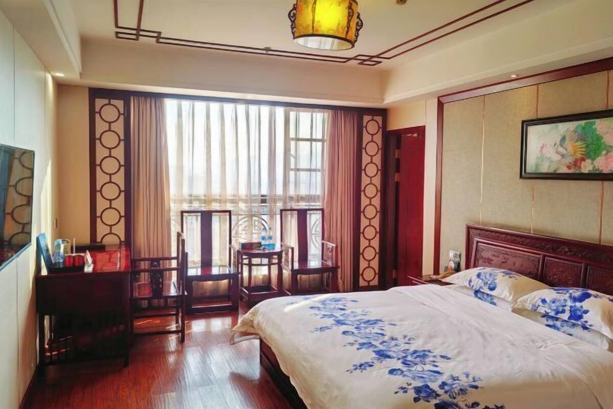 A hotel room with a bed, a desk, a chair, a window with curtains, and a painting on the wall.
