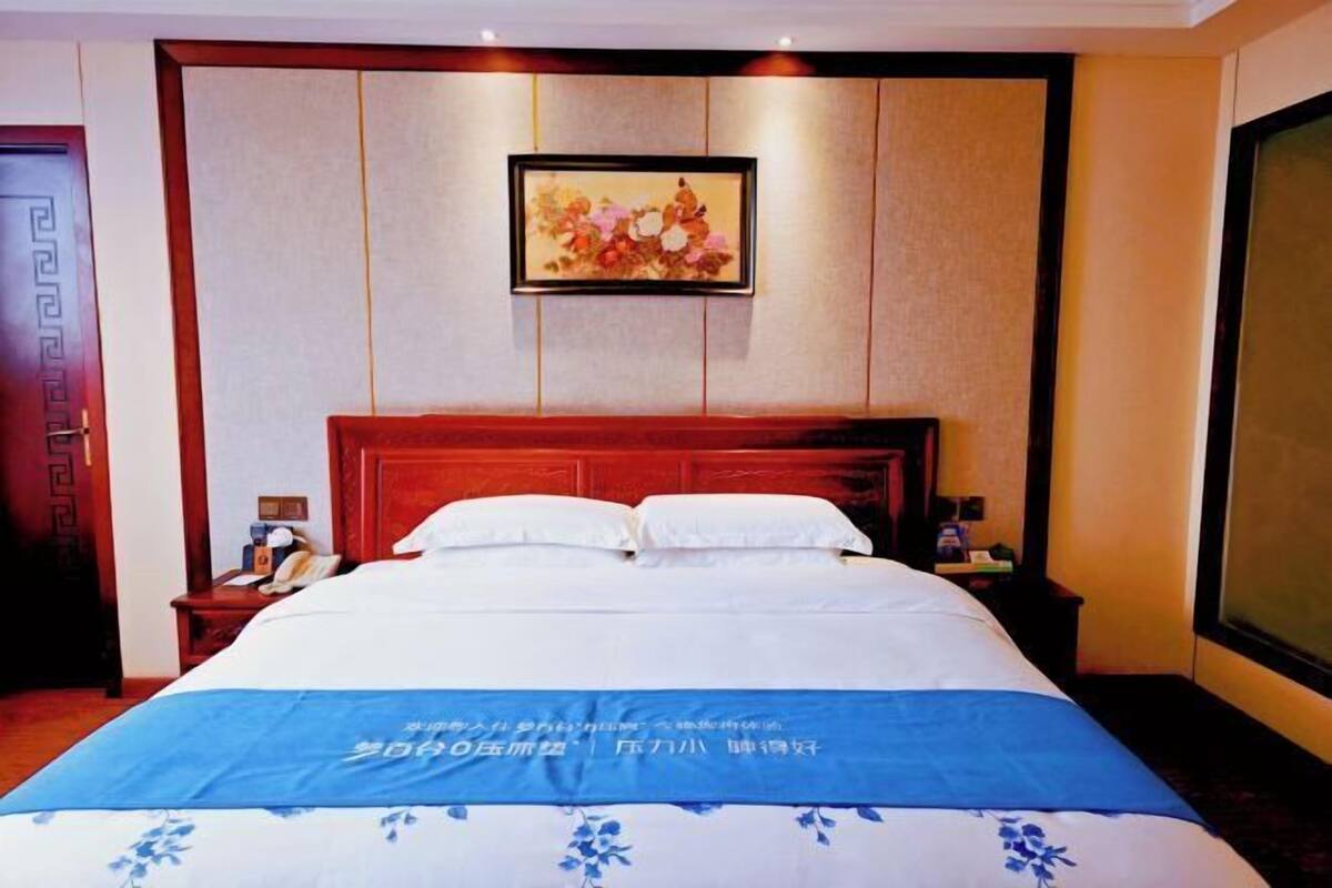 A hotel room with a large bed, a wooden headboard, a nightstand with a lamp, and a framed picture on the wall.