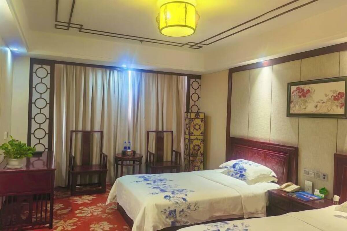A hotel room with two beds, a desk with a chair, a small table, and a potted plant.