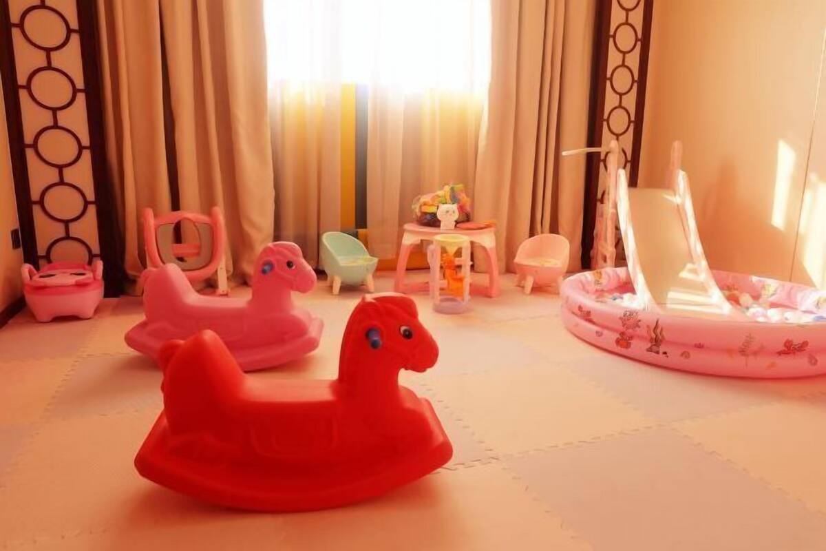 A room with a play area including a red rocking horse, a pink rocking horse, and a pink chair. There is a small table with toys and a white chair with a floral pattern.