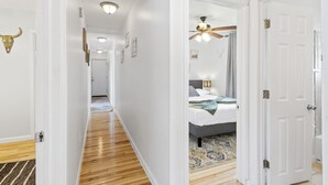A hallway with wooden flooring leading to a bedroom with a bed, a ceiling fan, and a patterned rug.