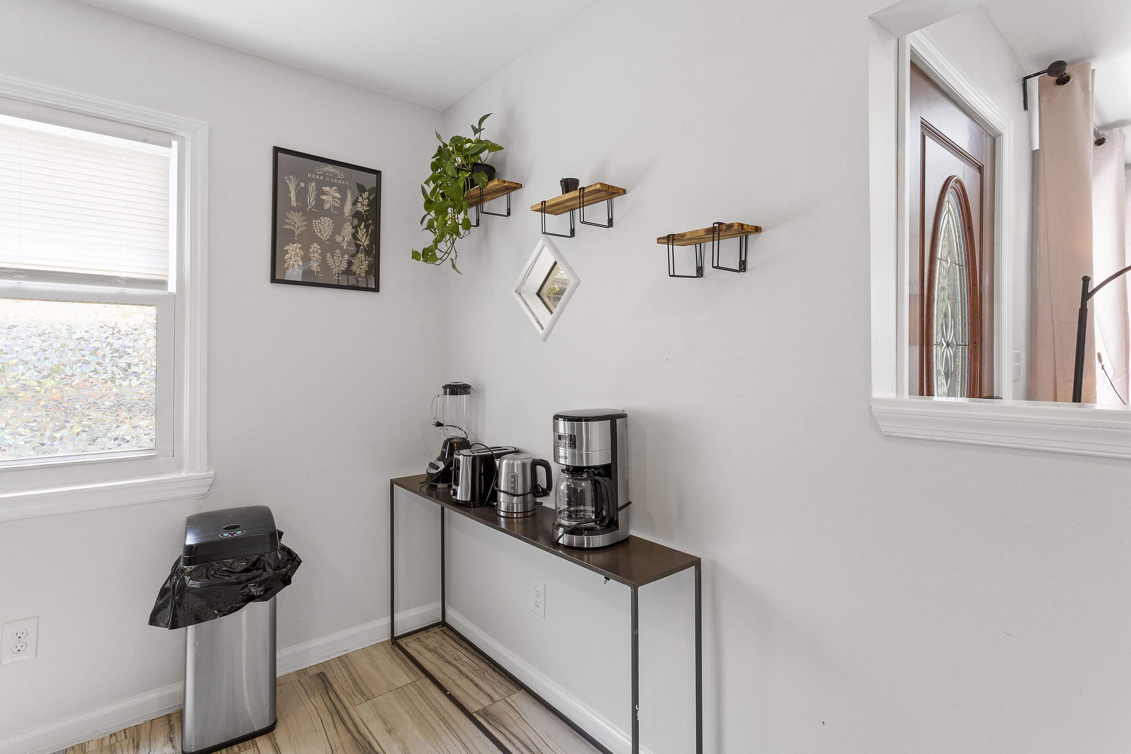 A compact kitchen with a coffee maker, toaster, and a trash bin. There are three wall-mounted shelves with plants and a framed botanical print.