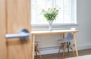 A room with a wooden table, two grey chairs, a white vase with flowers, and a window with blinds. - James' Place at The Taff (Merthyr Tydfil)