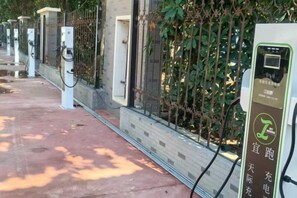 A row of electric vehicle charging stations along a paved walkway with a metal fence and greenery in the background.