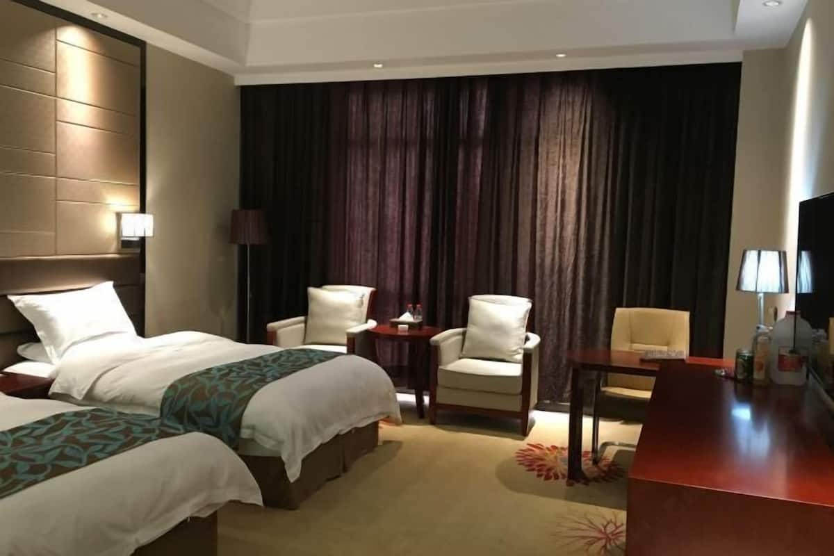 A hotel room with two beds, a desk, two chairs, a TV, and a large window with curtains.