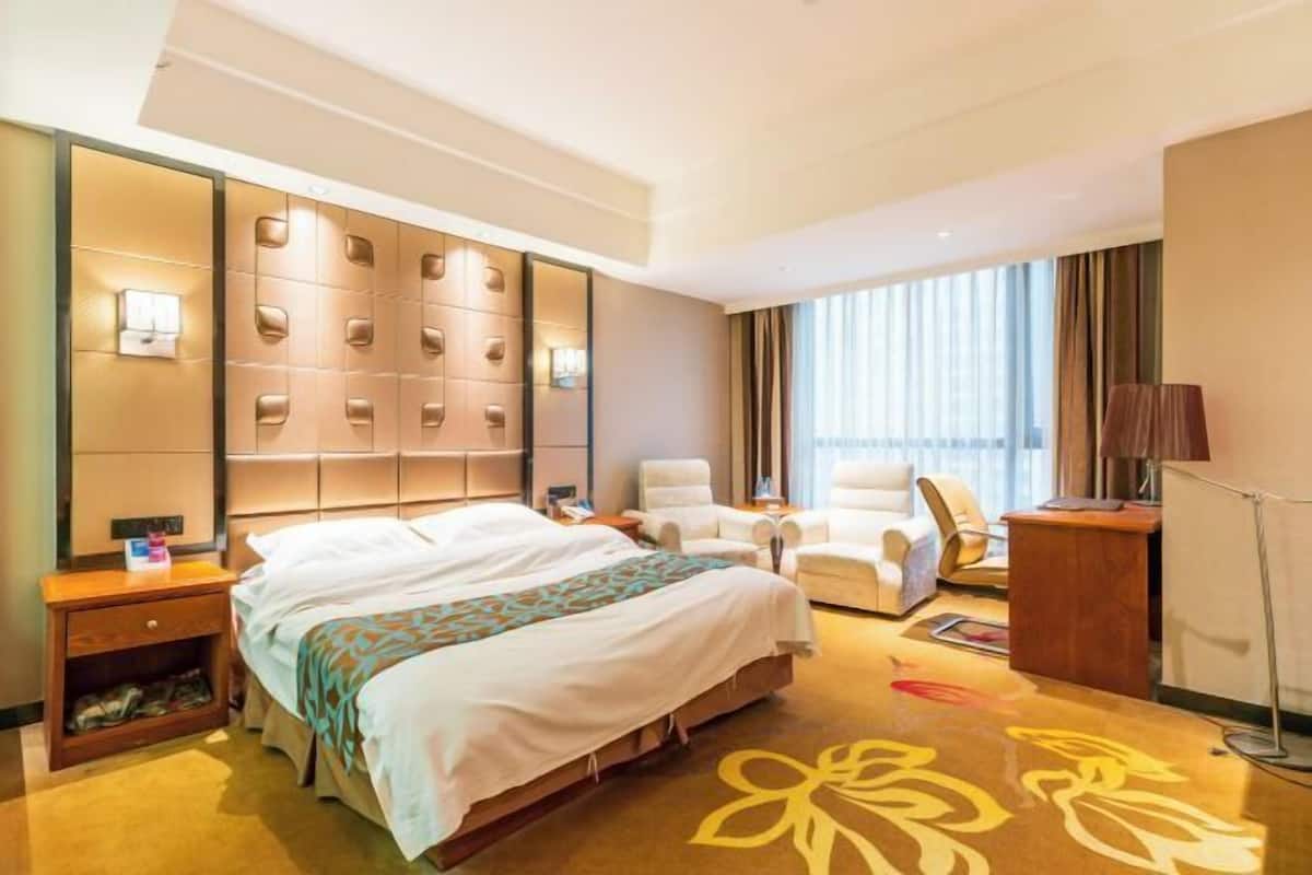 A hotel room with a large bed, a seating area with chairs, a desk, and a large window with curtains.
