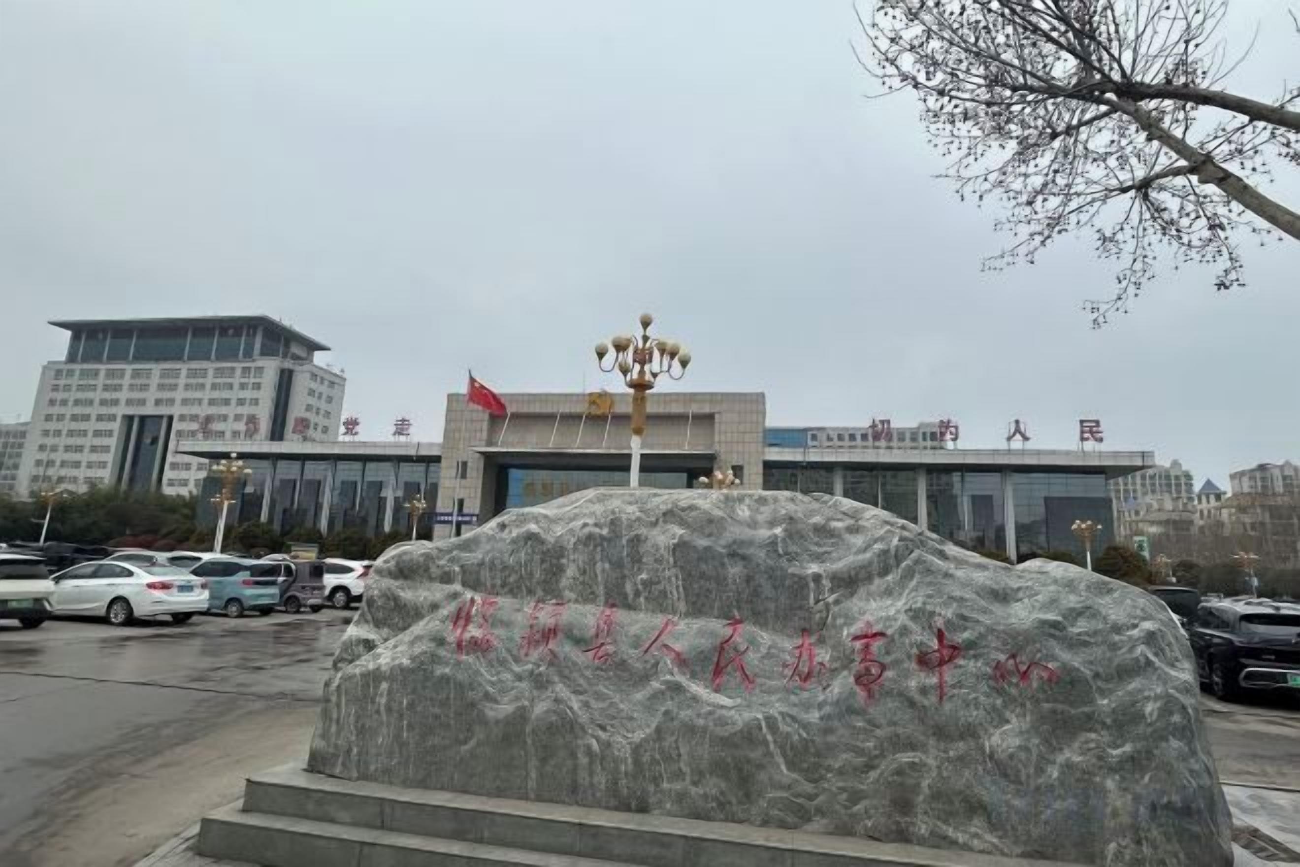 A stone monument with inscriptions, a modern building with a flag, and parked cars in a parking lot.