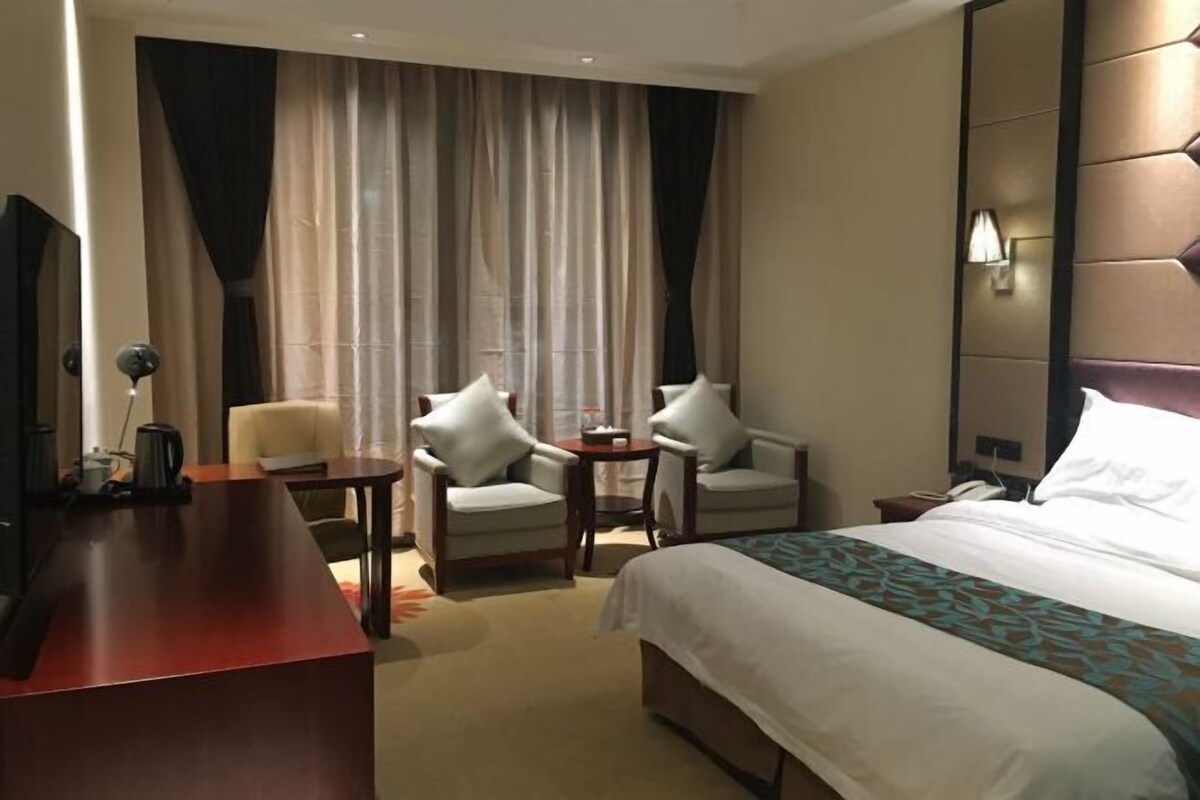 Quarto de hotel com cama, duas poltronas, escrivaninha, televisão e uma janela grande com cortinas.