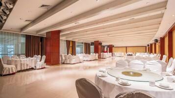 Un salón de banquetes con mesas redondas dispuestas para comer, sillas blancas y un techo alto con vigas de madera.