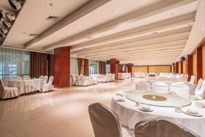 A banquet hall with round tables set for dining, white chairs, and a high ceiling with wooden beams.