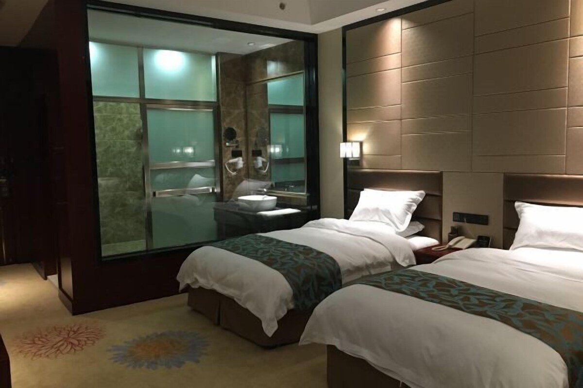 Quarto de hotel moderno com duas camas, uma janela grande, banheiro com box de vidro e um tapete decorativo.