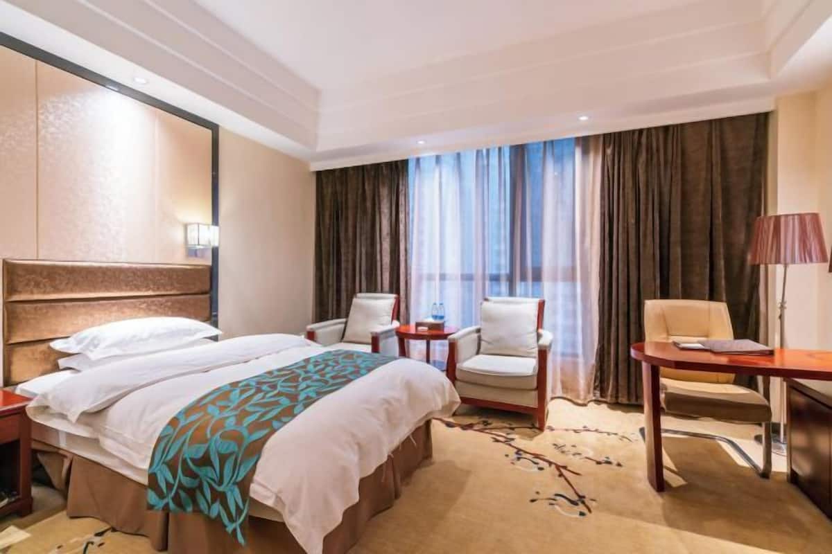 A hotel room with a bed, two chairs, a table, a lamp, and a view of the outside through large curtains.
