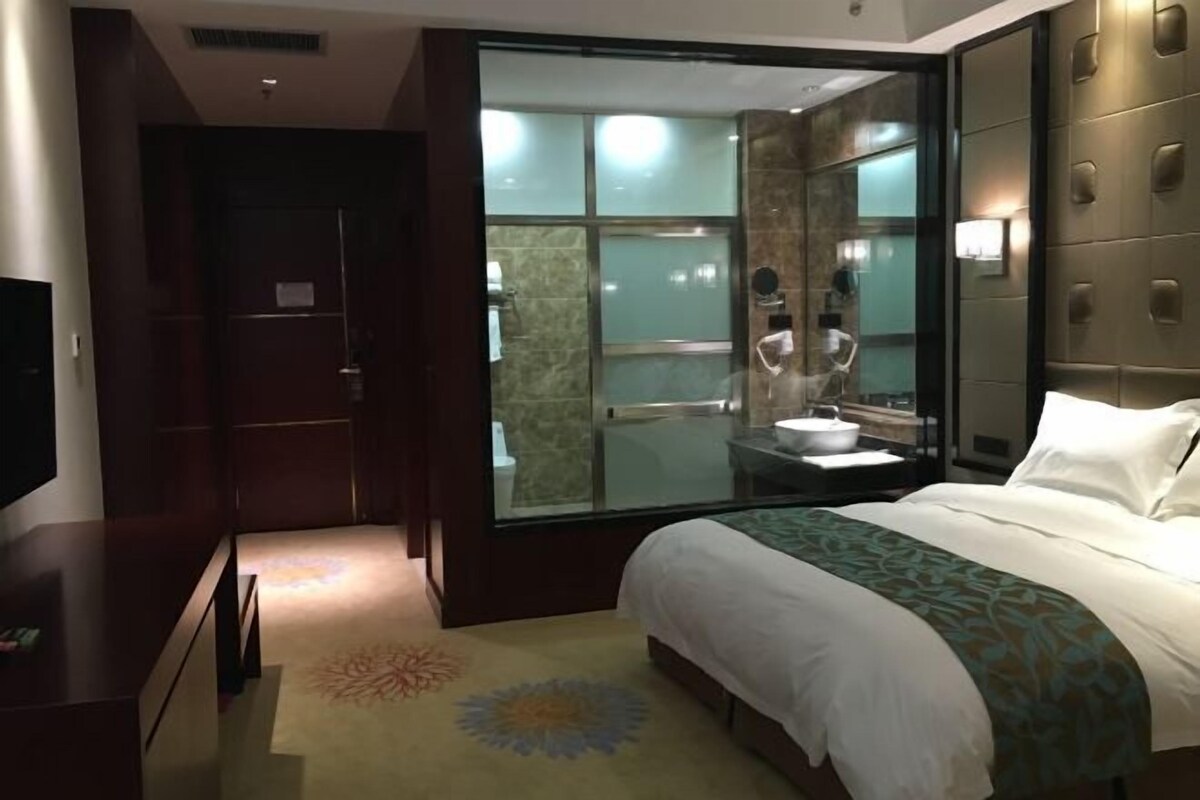 Quarto de hotel com uma cama grande, televisão, banheiro com box de vidro e vista para o banheiro através da porta de vidro.