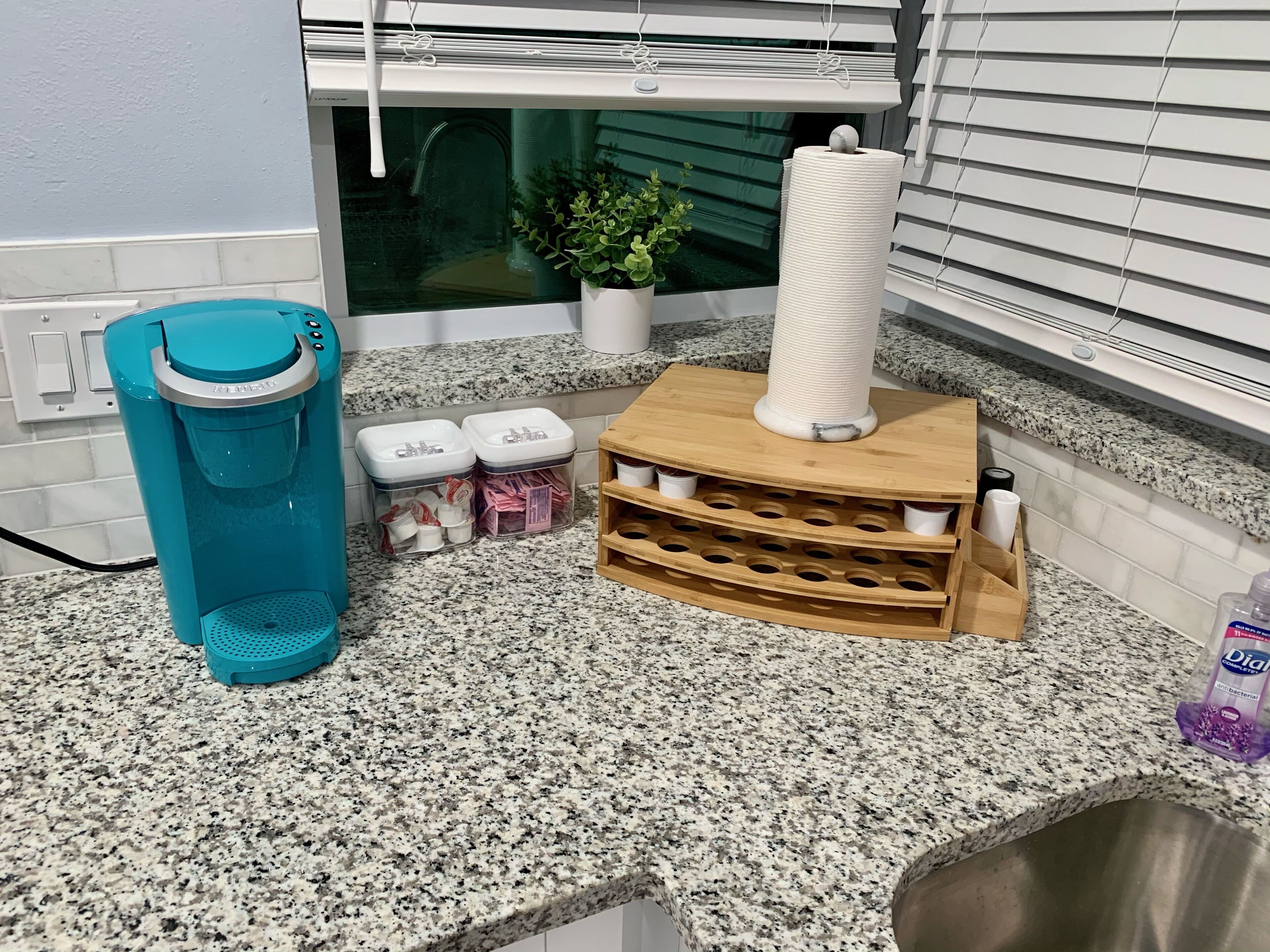Een keukenblad van graniet, een blauwe waterdispenser, twee doorzichtige plastic bakken met spullen, een houten rek met bekers, een potplant en een raam met jaloezieën.