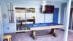 A pool table in a covered outdoor area with a ceiling fan, a television mounted on the wall, and a wooden stool. - Poolside Palms  (West Palm Beach)