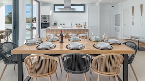 Une cuisine moderne dotée d’un îlot central, d’un coin repas avec une table en bois pouvant accueillir six personnes, et offrant une vue sur l’extérieur.