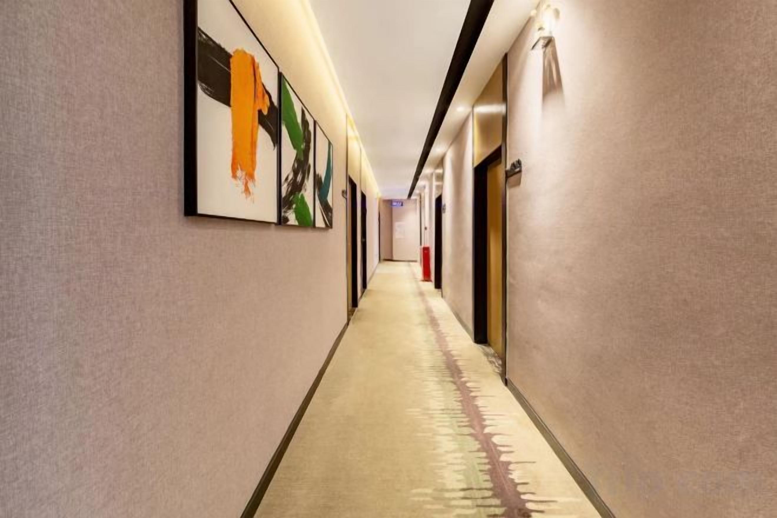 A long, narrow hotel corridor with framed artwork on the walls, a carpeted floor, and doors on the right side.