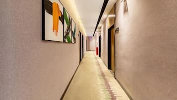 A long, narrow hotel corridor with framed artwork on the walls, a carpeted floor, and doors on the right side.