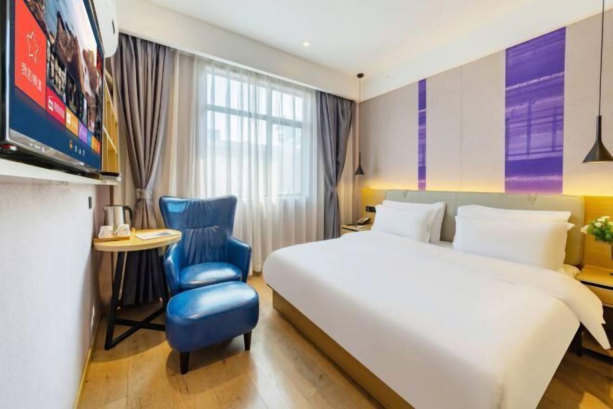 Habitación de hotel con una cama grande, un sillón azul, una mesita, un televisor y una ventana con cortinas.
